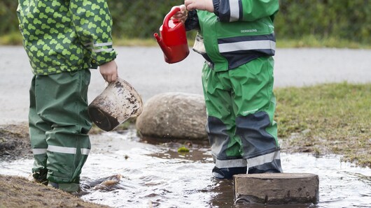 Bildet viser to barn i regntøy med vannkanne og bøtte som står i en sølepytt