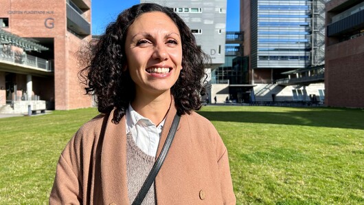 Bildet viser Rasha Abdallah, en smilende kvinne med krøllete mørkt hår og en brun kåpe. Hun står utendørs med grønt gress og noen moderne bygninger i bakgrunnen. Det er solskinn og klar blå himmel.